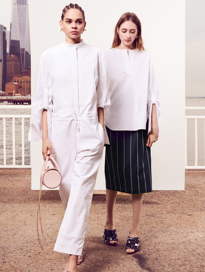 Two models pose in front of a white backdrop with the New York City skyline behind them. Holding a blush tube-like handbag, the model on the left is wearing a long-sleeve button-up white romper. The model on the right is wearing white long-sleeve V-neck blouse and navy and a white pinstripe skirt.