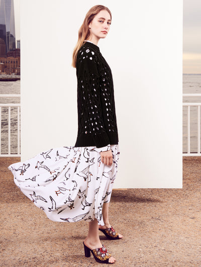 The model poses in front of a white backdrop with the New York City skyline behind her, wearing a black cable knit pullover layered over a silk long-sleeve dress printed with an all-over bird design.