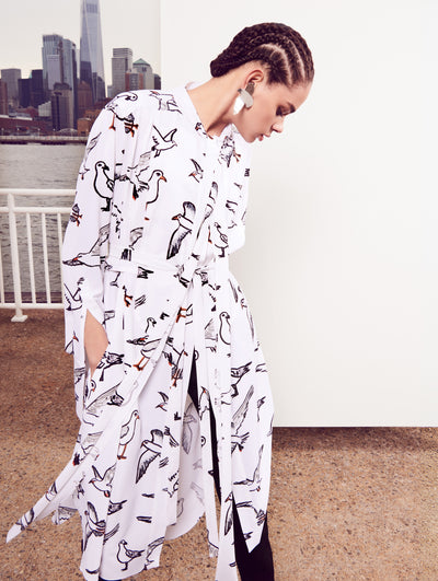 The model poses in front of a white backdrop with the New York City skyline behind her, wearing a silk long-sleeve tie-neck tunic printed with an all-over bird design and black trousers.