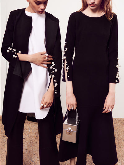 Two models pose in front of a white backdrop. The model on the left is wearing a black open-front coat with long split sleeves adorned with statement pearls layered over a white blouse and black trouser. Holding a silver cube-like handbag, the model on the right is wearing a black midi dress with three-quarter-length split sleeves adorned with statement pearls.