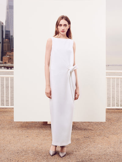 The model poses in front of a white backdrop with the New York City skyline behind her, wearing a white sleeveless column gown with a bow detail at the waist.