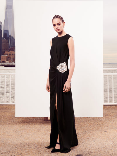 The model poses in front of a white backdrop with the New York City skyline behind her, wearing a black sleeveless draped evening gown with embroidered crystal detailing clustered at the waist.