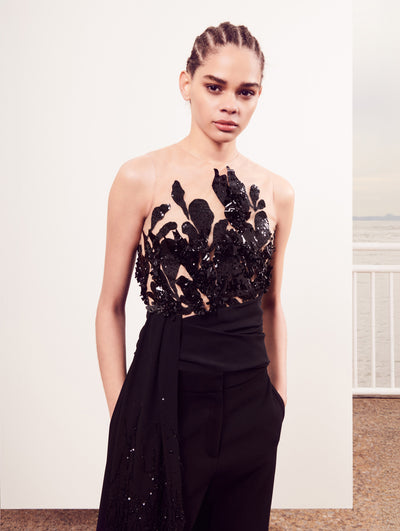 The model poses in front of a white backdrop, wearing an illusion-neck blouse with black floral sequin embroidery with a sash at the waist and black trousers.