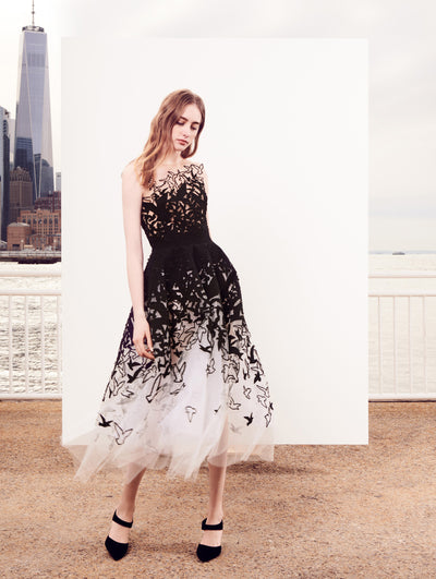 The model poses in front of a white backdrop with the New York City skyline behind her, wearing a white illusion-neck tulle cocktail dress featuring a black bird design concentrated at the bodice and fading into a delicate scatter toward the skirt.