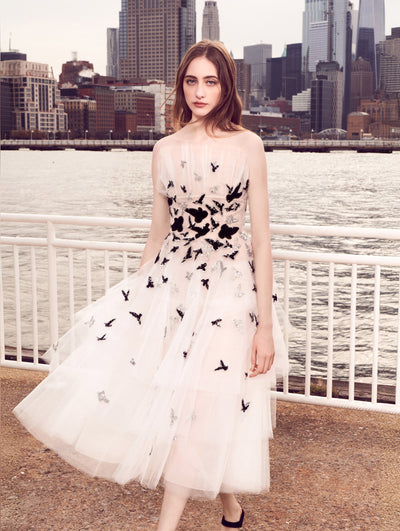The model poses in front of the New York City skyline, wearing a white strapless tulle cocktail dress embroidered with silver and black sequin birds concentrated at the bodice and fading into a delicate scatter toward the skirt.