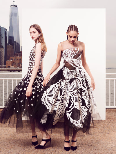 Two models pose in front of a white backdrop with the New York City skyline behind them. The model on the left is wearing a sleeveless illusion-neck black tulle cocktail dress decorated with and all-over white starfish design. The model on the right is wearing a sleeveless illusion-neck black tulle cocktail dress decorated with an all-over fish skeleton design.