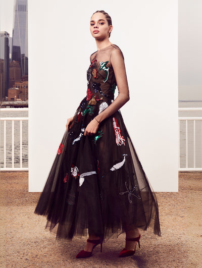 The model poses in front of a white backdrop with the New York City skyline behind her, wearing a long-sleeve illusion-neck black tulle evening gown embroidered with multi-colored sequin fish.