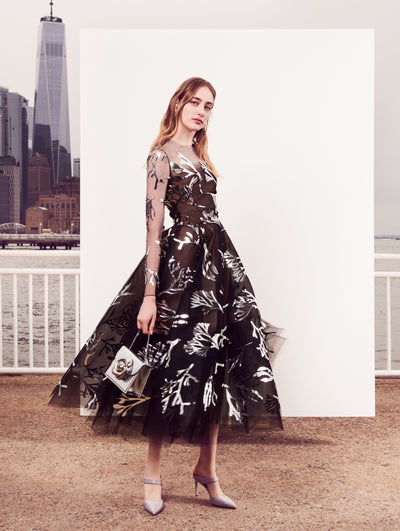 The model poses in front of a white backdrop with the New York City skyline behind her, wearing a long-sleeve illusion-neck black tulle cocktail dress printed with a silver all-over coral design. 