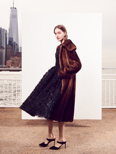 The model poses in front of a white backdrop with the New York City skyline behind her, wearing an open-front brown fur coat layered over a shimmering black cocktail dress.