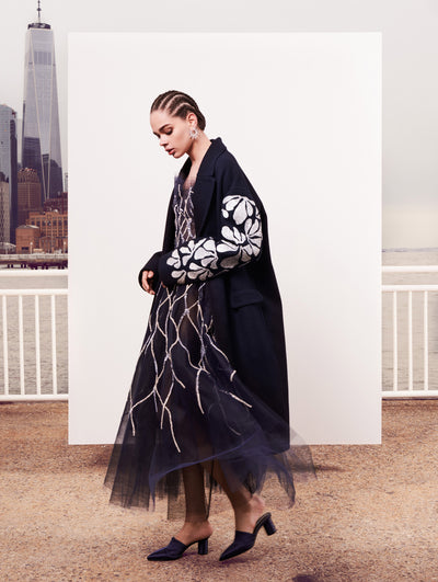 The model poses in front of a white backdrop with the New York City skyline behind her, wearing an open-front navy wool coat with white coral threadwork embroidery on the sleeves layered over a semi-sheer navy tulle cocktail dress embroidered with sinuous silver sequin coral.
