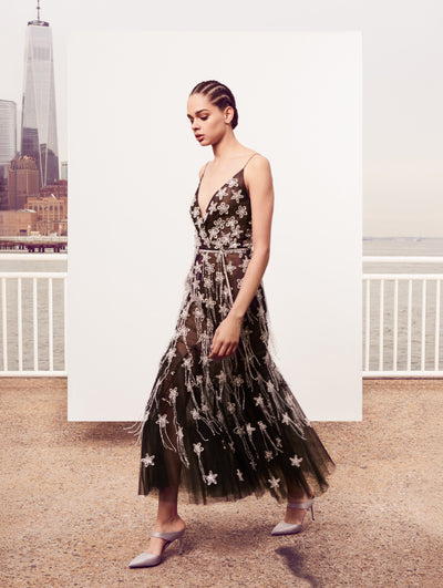The model poses in front of a white backdrop with the New York City skyline behind her, wearing a sleeveless semi-sheer black tulle evening gown with silver sequin star embroidery and bead fringe detailing.