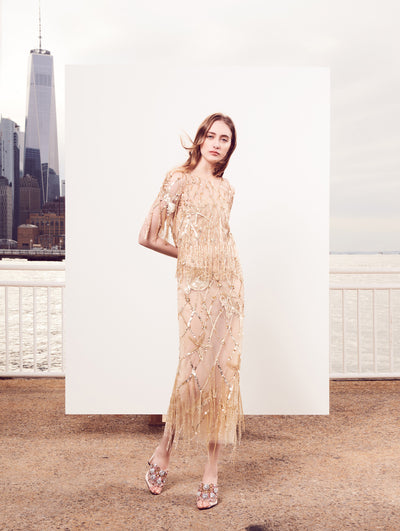The model poses in front of a white backdrop with the New York City skyline behind her, wearing a short-sleeve semi-sheer gold tulle evening gown with tonal sequin grid and starfish embroidery and bead fringe detailing.