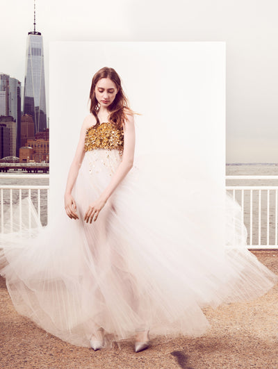 The model poses in front of a white backdrop with the New York City skyline behind her, wearing a strapless pleated tulle evening gown in bright white with a bodice made of gold sequin embroidery.