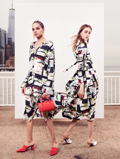 Two models pose in front of a white backdrop with the New York City skyline behind them. Holding a red tube-like handbag, the model on the left is wearing a multi-colored silk button-up long-sleeve blouse printed with postcards and a coordinating silk skirt printed with postcards. The model on the right is wearing a multi-colored silk button-up long-sleeve dress printed with postcards. 