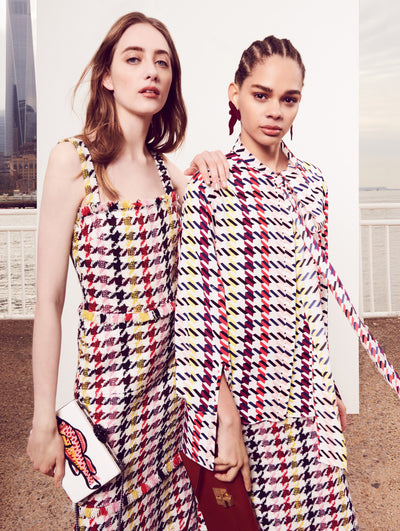 Two models pose in front of a white backdrop with the New York City skyline behind them. Holding a white clutch embroidered with a sequin fish, the model on the left is wearing a sleeveless multi-colored checked tweed dress with tiered fringe. Holding a red clutch with push-lock closure, the model on the right is wearing a silk long-sleeve tie-neck blouse printed with a multi-colored checked design and coordinating multi-colored checked tweed skirt.