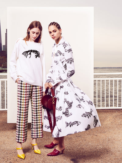 Two models pose in front of a white backdrop with the New York City skyline behind them. The model on the left is wearing a long sleeve knit pullover embroidered with a black sequin fish skeleton and multi-colored checked tweed trousers. Holding a red velvet tube-like handbag, the model on the right is wearing a white silk long-sleeve tie-neck dress printed with a black all-over fish skeleton design.
