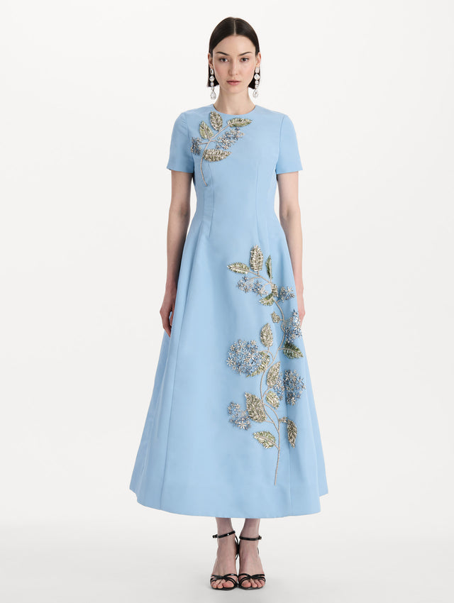 The model is wearing the Crystal Flower Embroidered Faille Dress in the color light blue with short sleeves, a sculpted skirt, and crystal-embroidered floral motifs. Front View.
