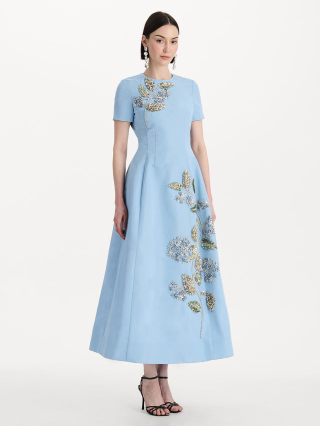 The model is wearing the Crystal Flower Embroidered Faille Dress in the color light blue with short sleeves, a sculpted skirt, and crystal-embroidered floral motifs. Side View.
