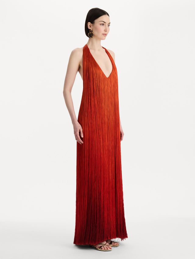 The model is wearing the Ombré Fringe Gown in Paprika Red. Featuring a deep, halter neckline, and full fringe silhouette. Side View.