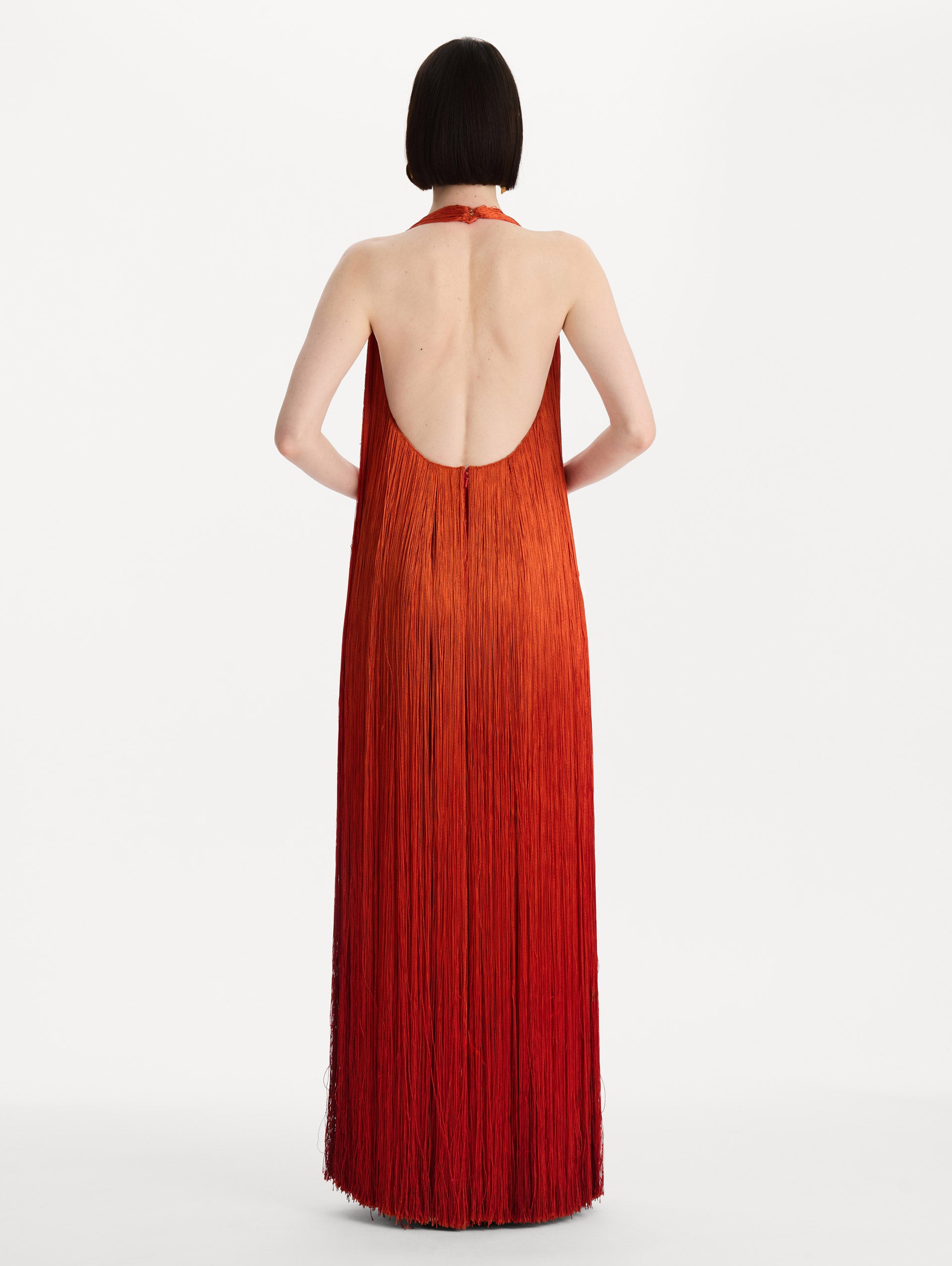 The model is wearing the Ombré Fringe Gown in Paprika Red. Featuring a deep, halter neckline, and full fringe silhouette. Back View.