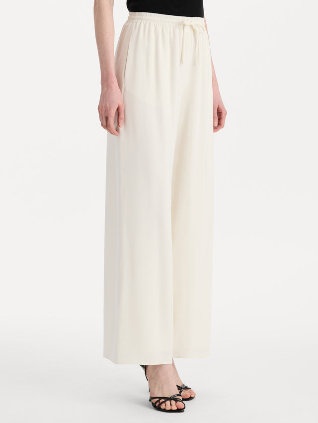 The model is wearing the Silk Georgette Drawstring Pant in the color ivory featuring straight, wide-legs and an elastic waistband finished with a drawstring tie. Side View.