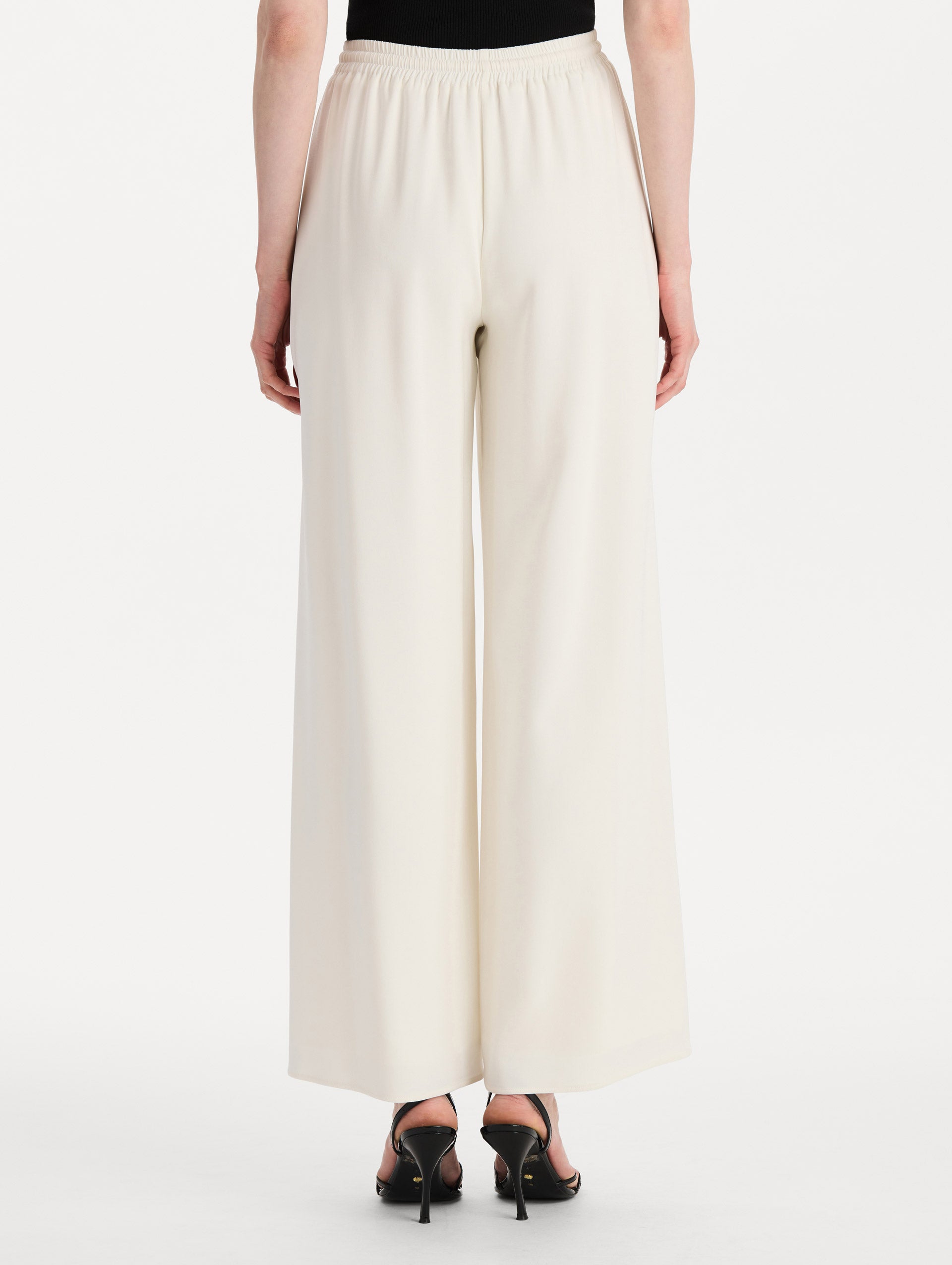 The model is wearing the Silk Georgette Drawstring Pant in the color ivory featuring straight, wide-legs and an elastic waistband finished with a drawstring tie. Back View.