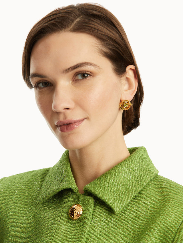 The model is wearing gold textured studs with a topaz crystal in the center, paired with a green jacket. Front view