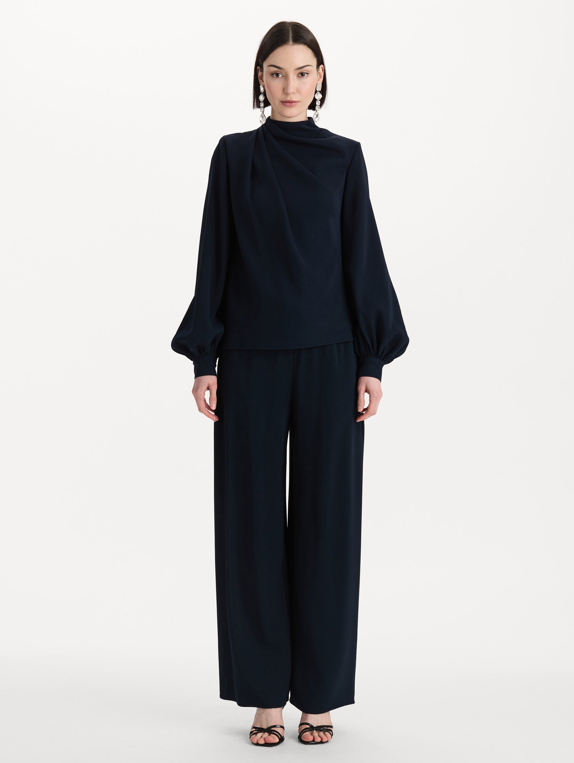 The model is wearing the Draped Georgette Blouse in the color Navy. Featuring silk georgette, a draping mock-neckline, and long, billowed sleeves. Front View.