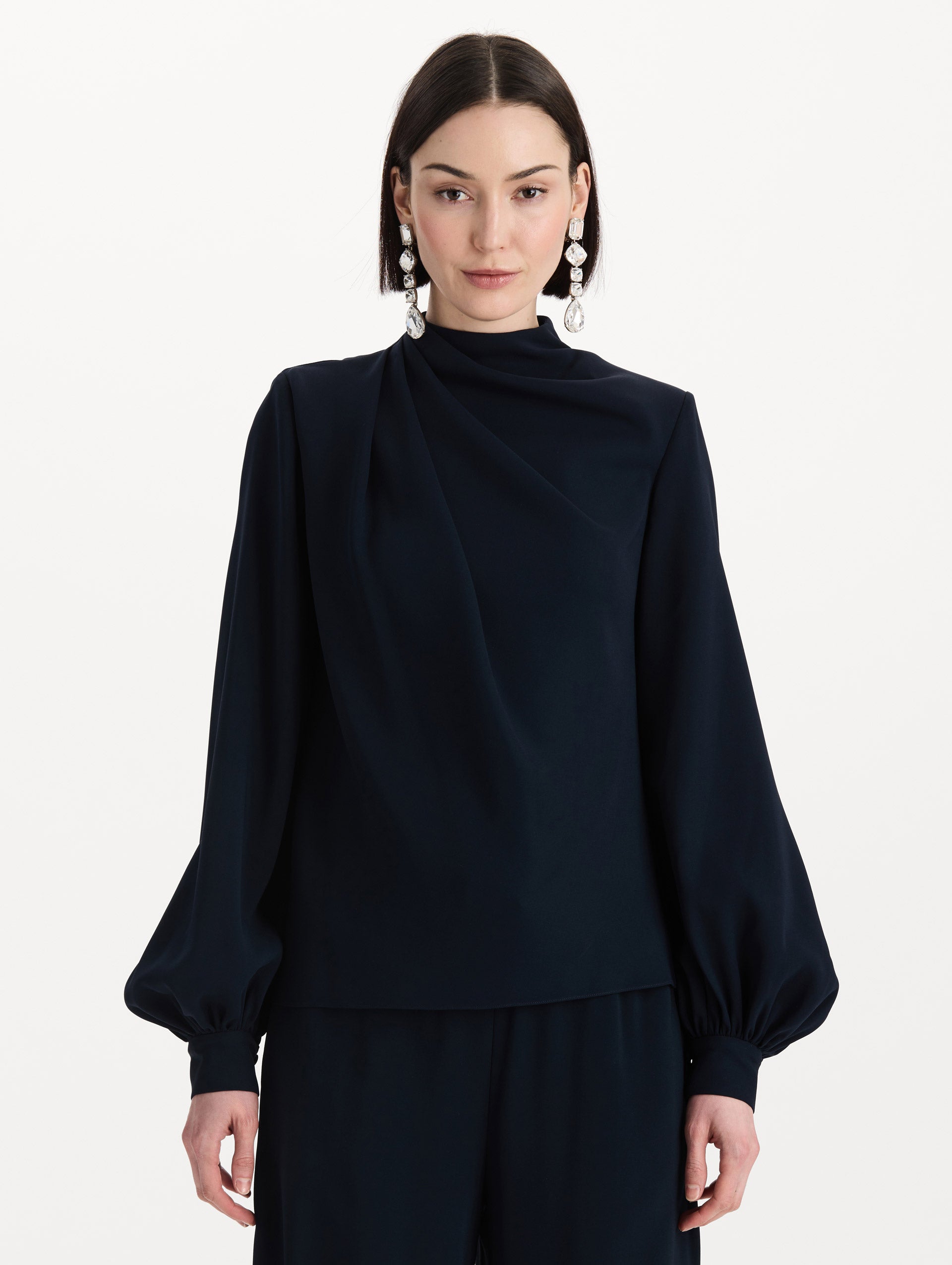 The model is wearing the Draped Georgette Blouse in the color Navy. Featuring silk georgette, a draping mock-neckline, and long, billowed sleeves. Front View.