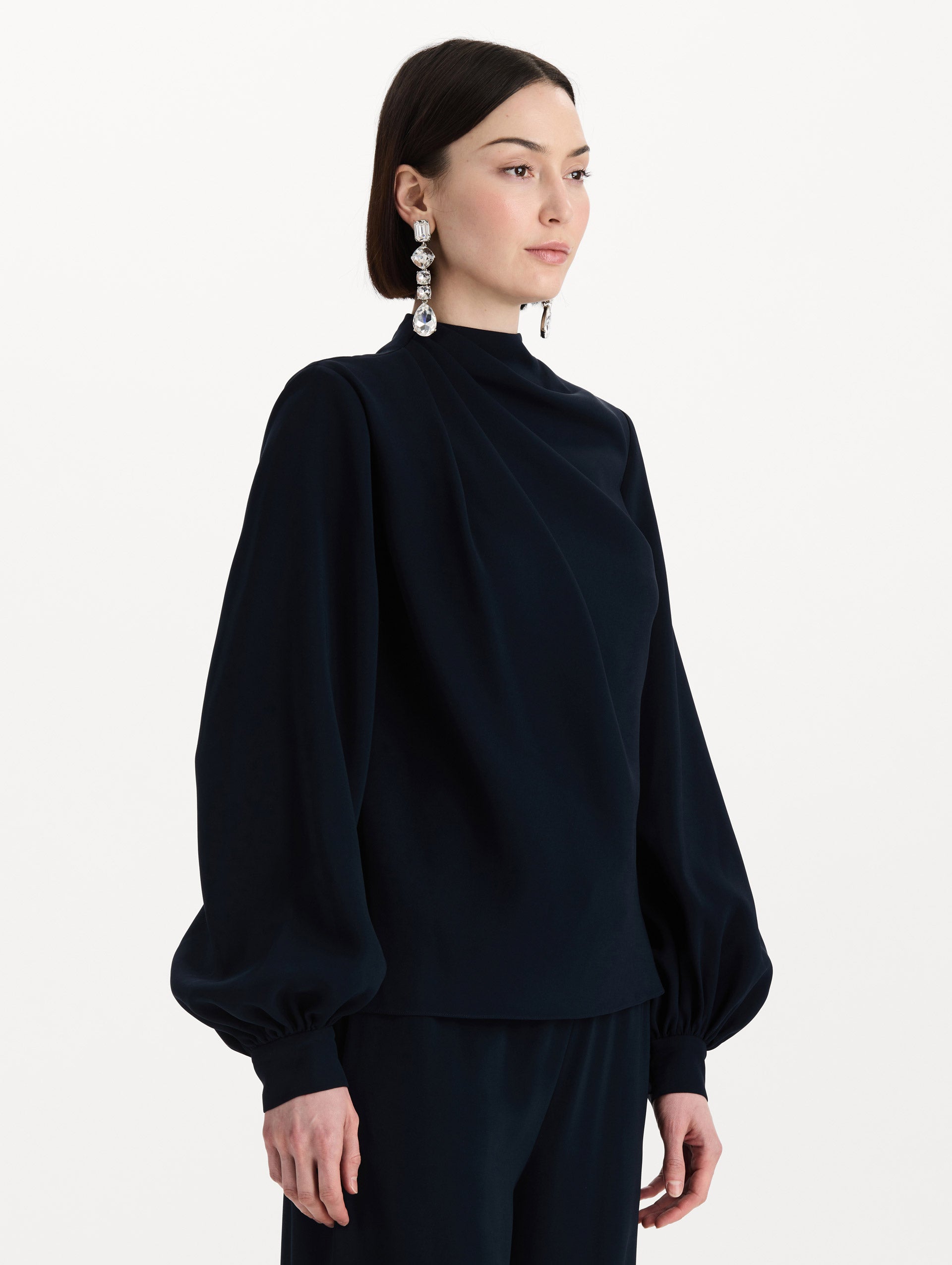 The model is wearing the Draped Georgette Blouse in the color Navy. Featuring silk georgette, a draping mock-neckline, and long, billowed sleeves. Side View.