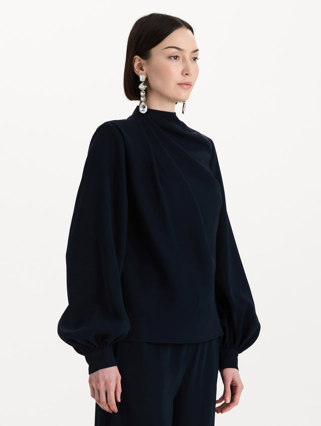 The model is wearing the Draped Georgette Blouse in the color Navy. Featuring silk georgette, a draping mock-neckline, and long, billowed sleeves. Side View.