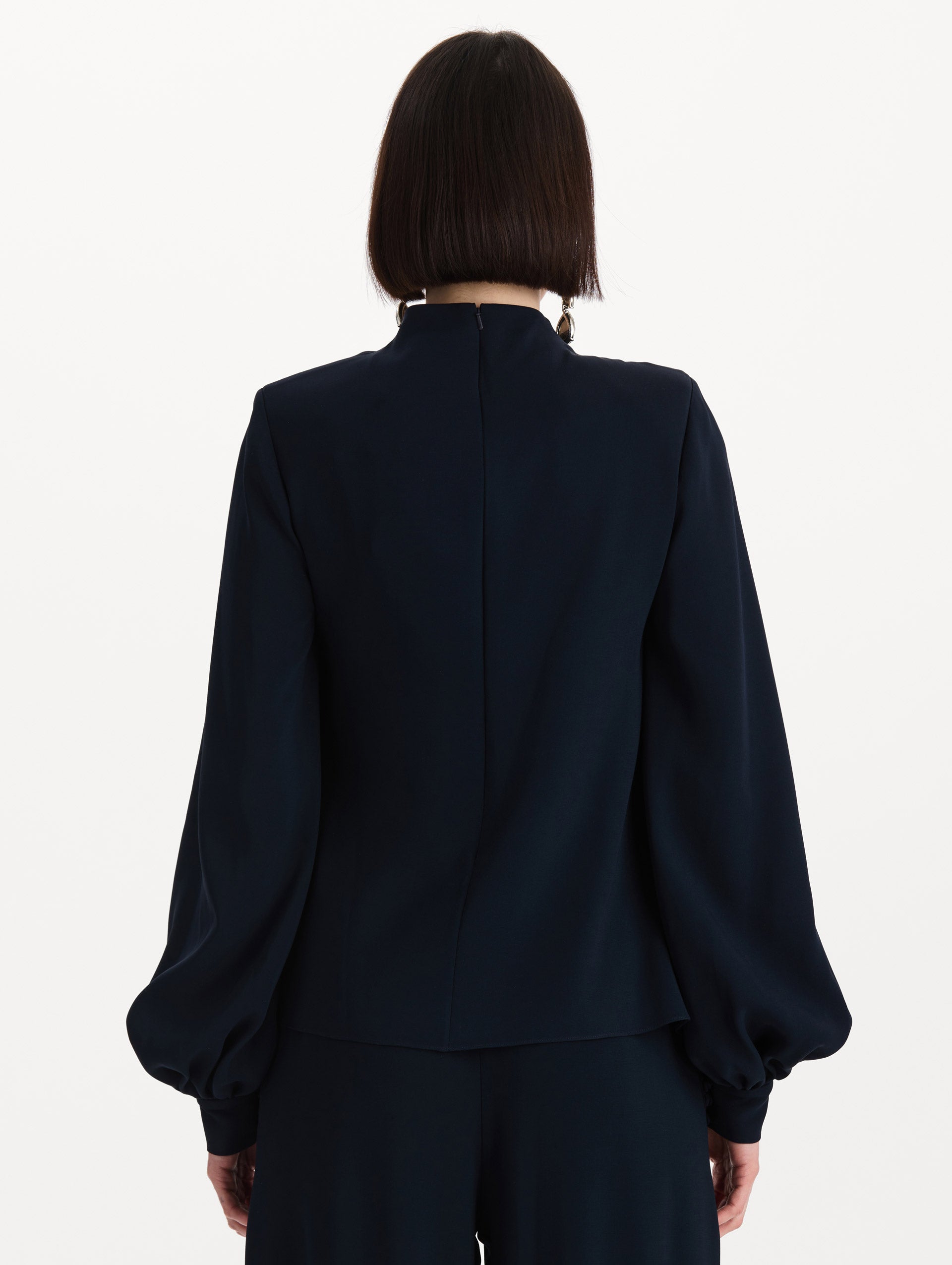 The model is wearing the Draped Georgette Blouse in the color Navy. Featuring silk georgette, a draping mock-neckline, and long, billowed sleeves. Back View.