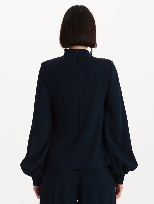 The model is wearing the Draped Georgette Blouse in the color Navy. Featuring silk georgette, a draping mock-neckline, and long, billowed sleeves. Back View.