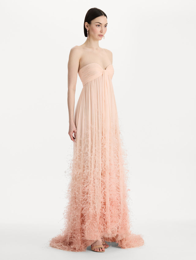 The model is wearing a strapless, sweetheart neckline gown in Blush Pink. Featuring a long flowing skirt with feather details. Side View.