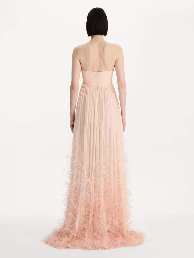 The model is wearing a strapless, sweetheart neckline gown in Blush Pink. Featuring a long flowing skirt with feather details. Back View.