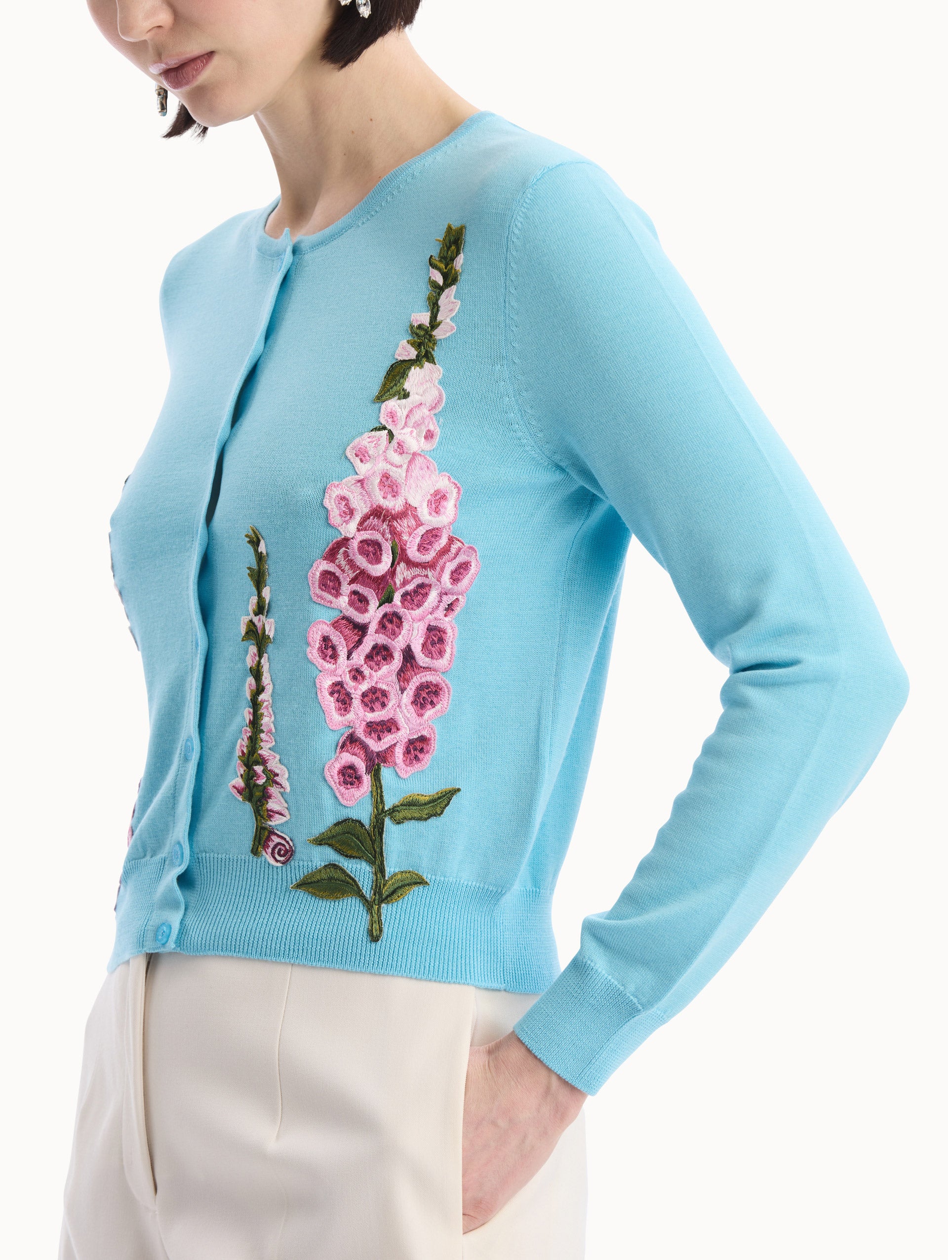 The model is wearing the Foxglove Threadwork Cardigan in vibrant light blue color with floral embroidery and buttons along the center. Side View.