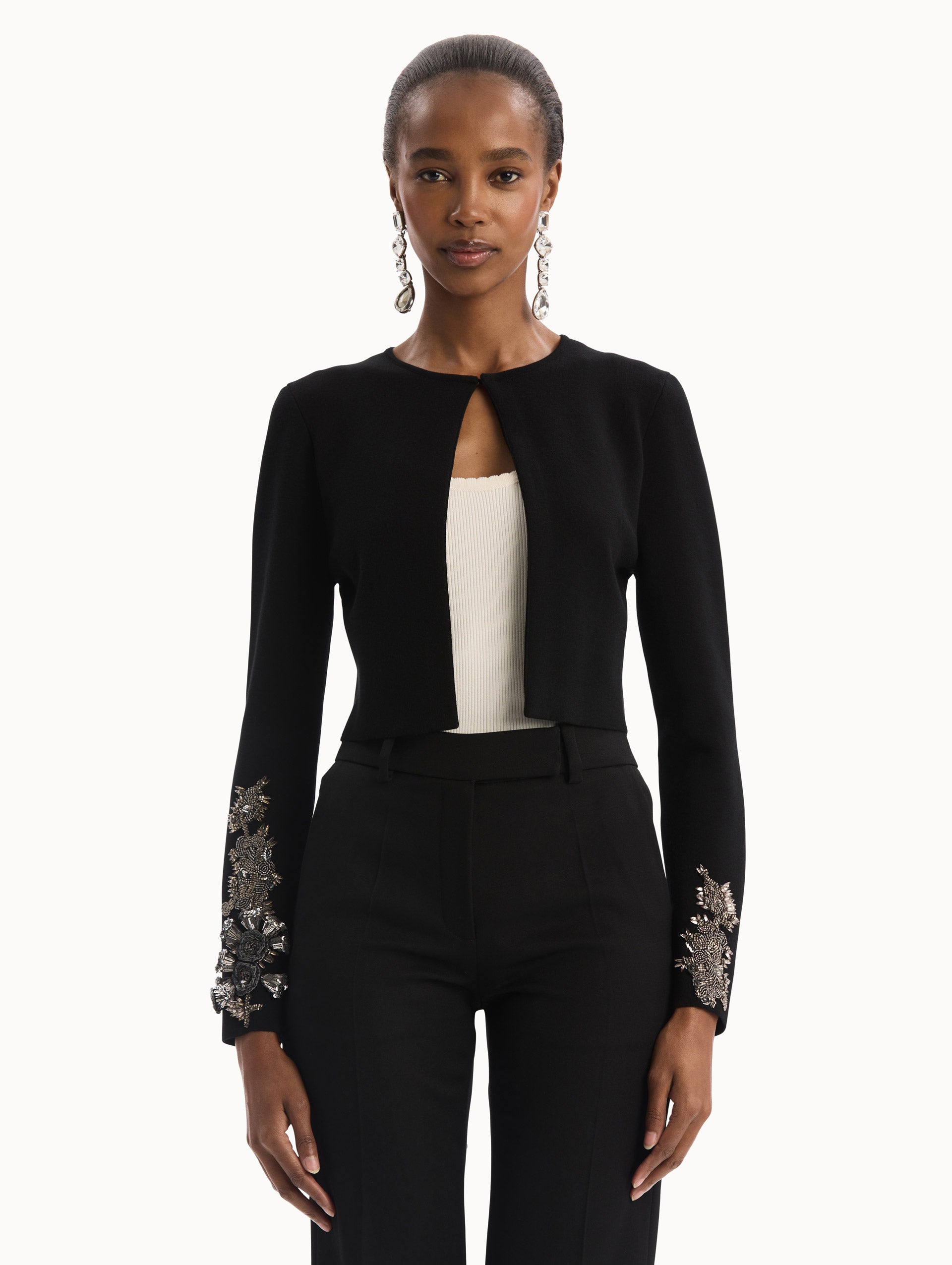 The model is wearing the Black Feather Sequin Embroidered Knit Cardigan featuring an open front, textured inset panel, and silver floral embroidery along the sleeves. Front View.