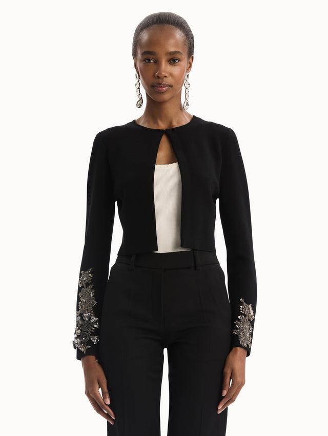 The model is wearing the Black Feather Sequin Embroidered Knit Cardigan featuring an open front, textured inset panel, and silver floral embroidery along the sleeves. Front View.