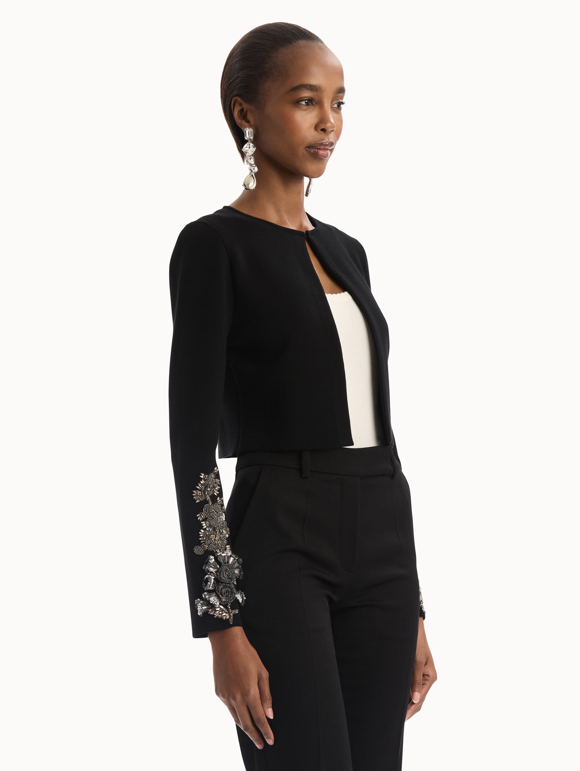 The model is wearing the Black Feather Sequin Embroidered Knit Cardigan featuring an open front, textured inset panel, and silver floral embroidery along the sleeves. Side View.