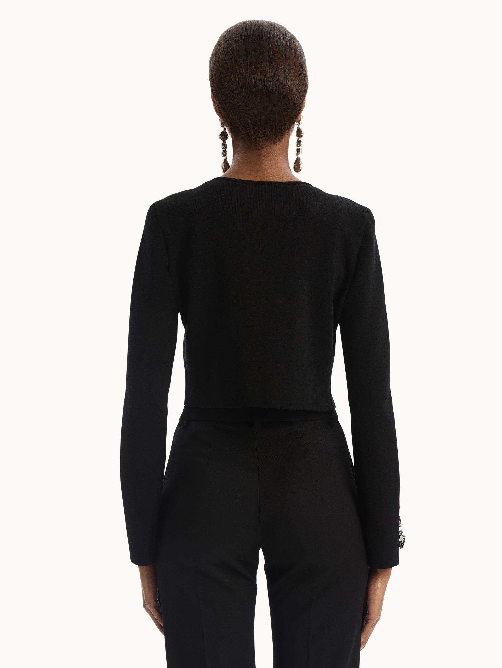 The model is wearing the Black Feather Sequin Embroidered Knit Cardigan featuring an open front, textured inset panel, and silver floral embroidery along the sleeves. Back View.