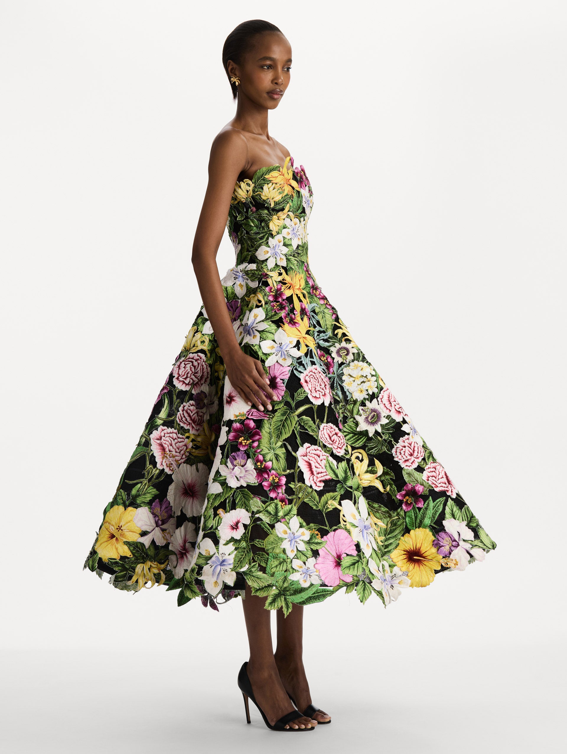 The model is wearing the Dominican Mixed-Floral Guipure Gown. Featuring a strapless neckline, fitted bodice, and full skirt. Side View.