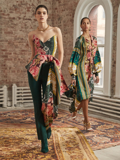 Two models pose in a loft with exposed brick walls and large arched windows behind them, standing on Persian rugs. The model on the left wears a strapless blouse with a multi-colored patchwork print and drape detailing, paired with green trousers. Holding a green gardenia appliqué handbag, the model on the right wears a long-sleeve silk dress printed with a multi-colored patchwork design.