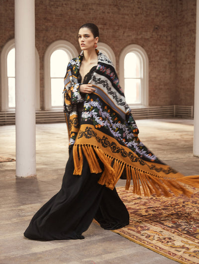 A model poses in a loft with exposed brick walls and large arched windows behind her, standing on a Persian rug. She wears an oversized shall with fringe trim and a multi-colored patchwork design, layered over a black jumpsuit with a V-neck lace bodice and wide-leg pants.