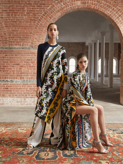 Two models pose in a loft with exposed brick walls and large arched windows behind them, standing on Persian rugs. The model on the left wears an oversized long-sleeve knit pullover with an asymmetrical hemline and multi-colored patchwork design, layered over black and white trousers with threadwork embroidered florals. Seated, the model on the right wears a long-sleeve silk evening gown printed with the same multi-colored patchwork design.