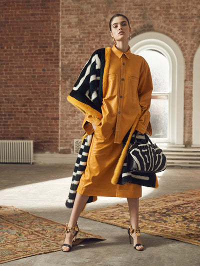 A model poses in a loft with exposed brick walls and large arched windows behind her, standing on a Persian rug. Holding a zebra print top-handle bag, the model wears a color-blocked orange, black and white fur shawl, layered over an orange long-sleeve button-up leather blouse, paired with a coordinating orange leather skirt.