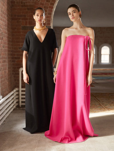 Two models pose in a loft with exposed brick walls, large arched windows and Persian rugs behind them. The model on the left wears a black short-sleeve V-neck wool caftan. The model on the right wears a bright pink strapless bow-detailed evening gown.