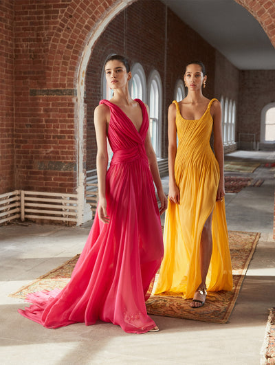 Two models pose in a loft with exposed brick walls and large arched windows behind them, standing on Persian rugs. The model on the left wears a pink sleeveless V-neck draped evening down. The model on the right wears an orange sleeveless scoop-neck draped evening down.
