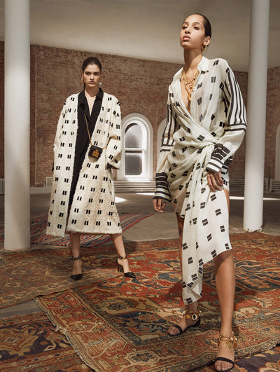 Two models pose in a loft with exposed brick walls and large arched windows behind them, standing on Persian rugs. The model on the left wears a white a black patterned open-front fur jacket, layered over a black button-up dress, accented with a crossbody micro black cube-like handbag. The model on the right wears a black and white patterned long-sleeve V-neck dress with an asymmetrical hemline.