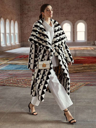 A model poses in a loft with exposed brick walls, large arched windows and Persian rugs behind her. She wears a long-sleeve spread-collar teddy coat with a black and white geometric circle motif, layered over a white button-up blouse and white trousers, accented with a white crossbody push-lock closure shoulder bag.