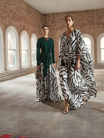 Two models pose in a loft with exposed brick walls and large arched windows behind them. Holding a zebra print gardenia appliqué handbag, the model on the left wears a green long-sleeve blouse with a tie detail, paired with a zebra print silk maxi skirt. The model on the right wears a silk zebra print V-neck caftan.
